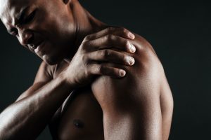 Cropped photo of young shirtless Black man with shoulder pain holding shoulder on black background; blog: signs of frozen shoulder
