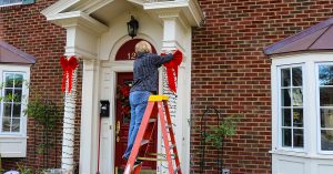 Woman on ladder putting Christmas decorations up on pillars of entrance of two story brick house with bay windows; blog: 9 Tips to Prevent Common Holiday Injuries
