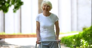 Cheerful senior woman with walking frame looking at camera outdoors, rehab; blog: What is the Best Age for Knee Replacement Surgery?