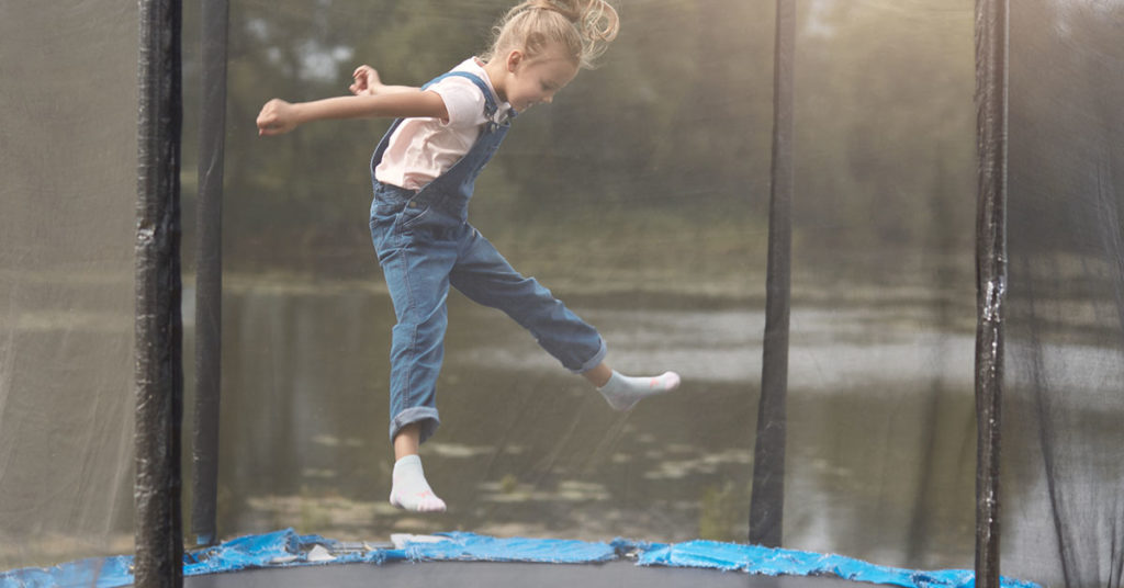 Full length photo of happy girl jumping high on trampoline in park, blonde female chicd with ponytail wearing white shirt and denim overalls, kid spending time in open air, having fun outdoor; blog: 9 Trampoline Safety Tip to Prevent Injuries