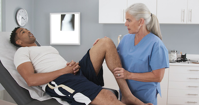 Patient at clinic with injured knee assisted by senior doctor. Elderly female doctor assessing mans leg injury; blog: benefits of orthopedic physical therapy